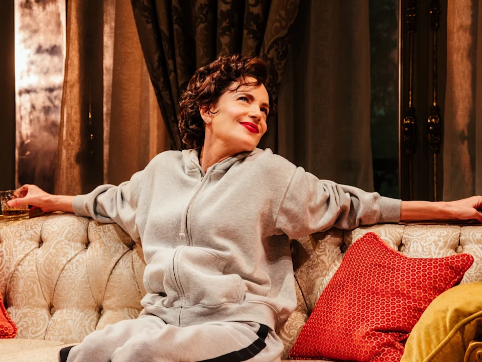 A person in a gray tracksuit sits relaxed on a patterned sofa, holding a drink and smiling slightly, with red cushions on either side.