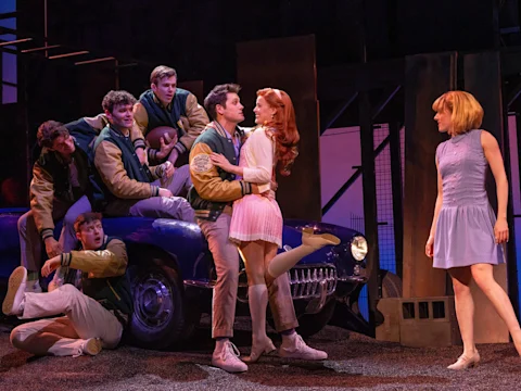 Actors on stage perform a dramatic scene; a group of young men in letterman jackets gather around a blue car while a man interacts with two women under theatrical lighting.