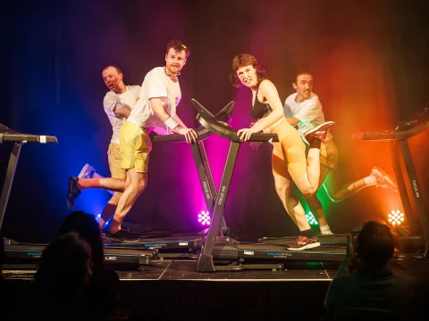 Four people in athletic wear perform energetically on treadmills, illuminated by colorful stage lights.