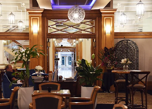 Hotel lobby with wooden furnishings, potted plants, round tables with chairs, and ornate lighting, with a hallway visible in the background.