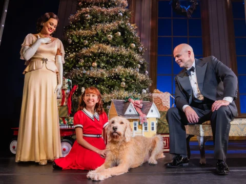 Three people and a dog are posed in front of a decorated Christmas tree. A woman in a peach dress stands; a girl in a red dress and a man in a tuxedo sit near the dog and a toy house.