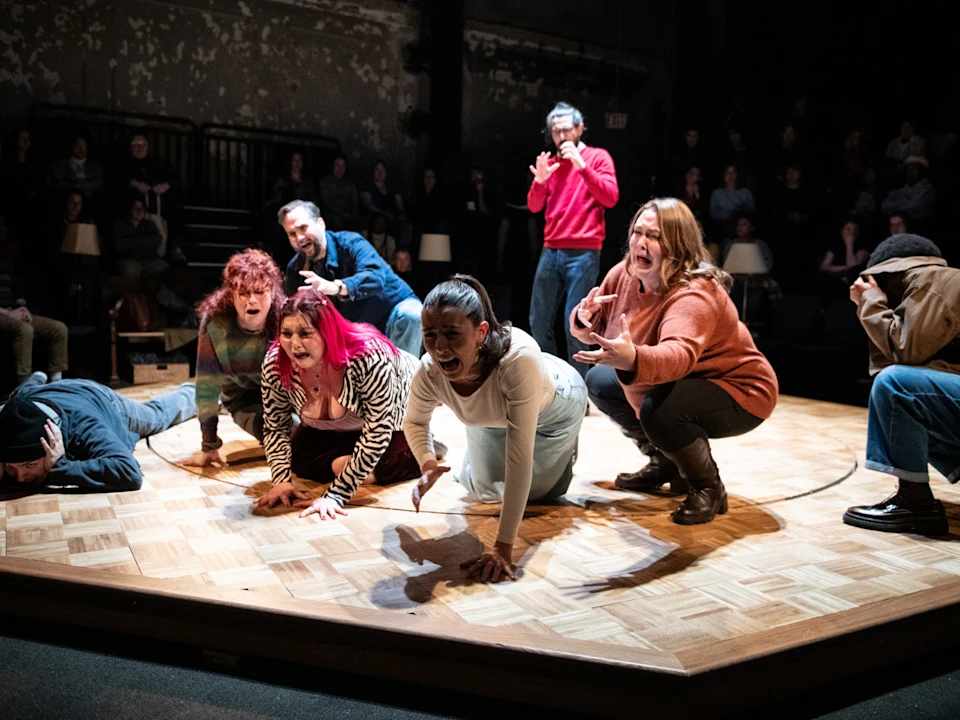 Seven actors on a wooden stage express intense emotions, some kneeling or crouching, while an audience watches in the background.