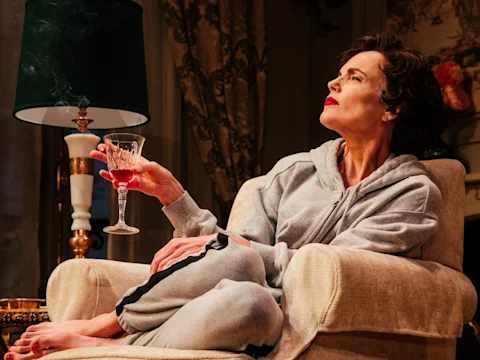 A woman in a gray tracksuit sits on an armchair, holding a glass of red wine and looking to the side, with a lamp and ornate decor in the background.