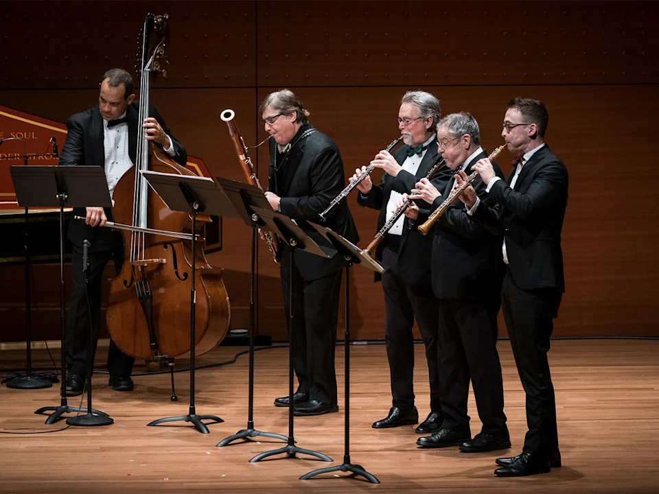 Five male musicians in formal wear perform on stage, playing double bass, bassoon, and three wind instruments, with music stands in front of them.