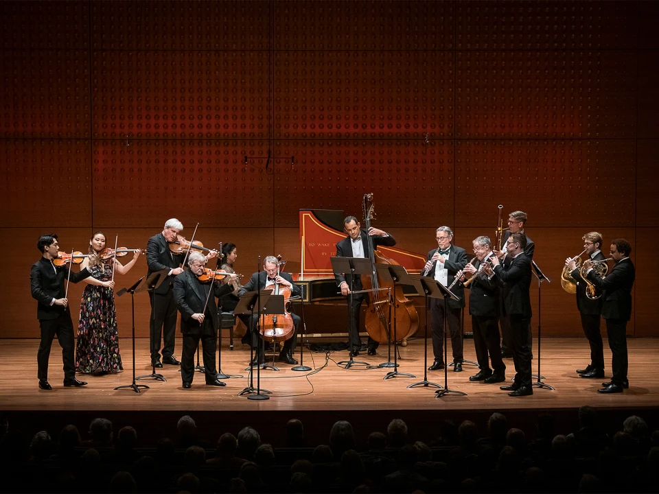 A group of musicians performs on stage with string, wind, and brass instruments. A harpsichord and a double bass are also visible. An audience watches from the foreground.