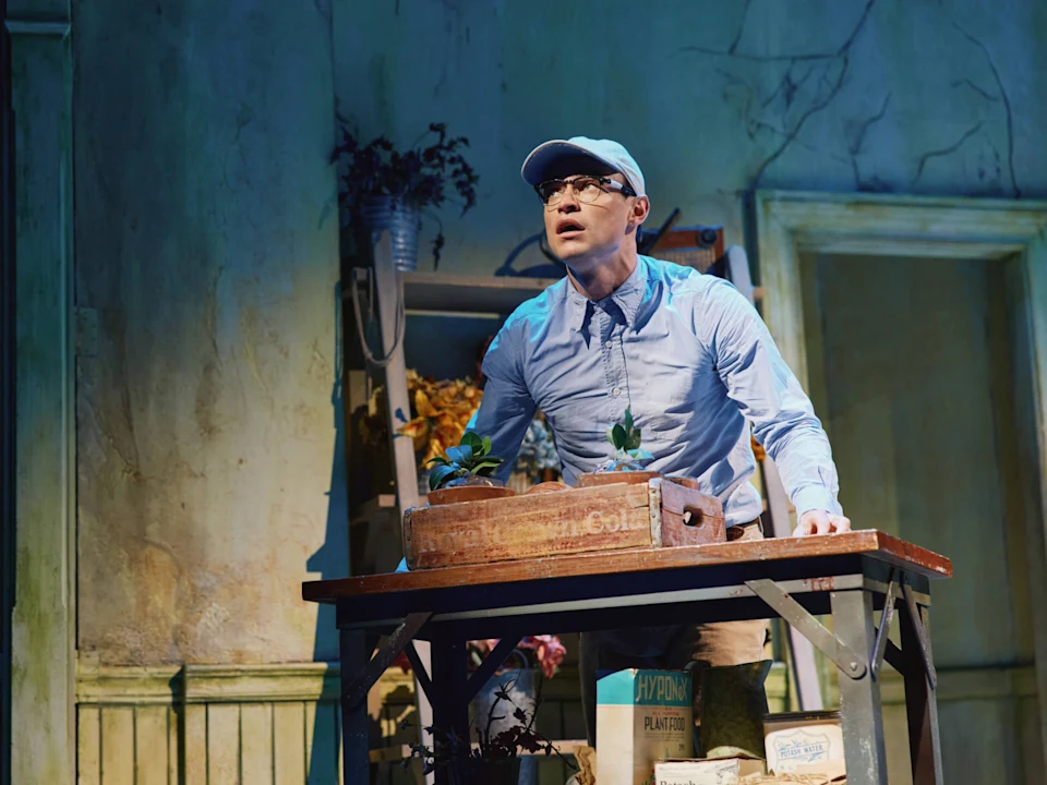A person in a light blue shirt, tie, and cap stands behind a table with plants and gardening supplies in a worn, indoor setting.