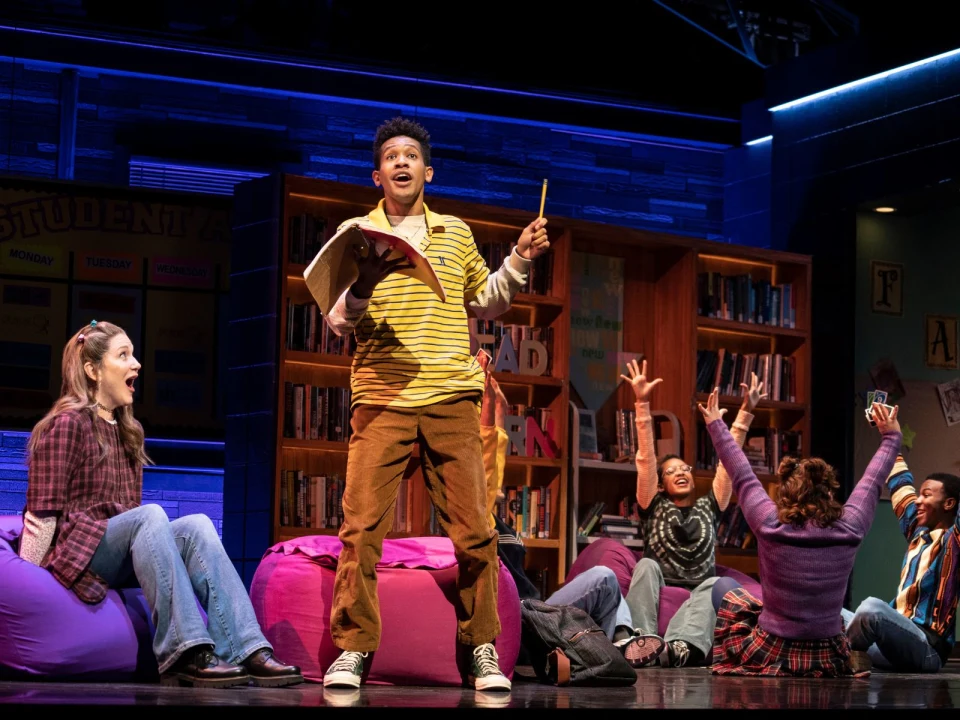 A group of young people on stage, some sitting on bean bags, one standing and holding a book. Others are reaching up or reacting animatedly. Shelves with books and educational decorations are in the background.