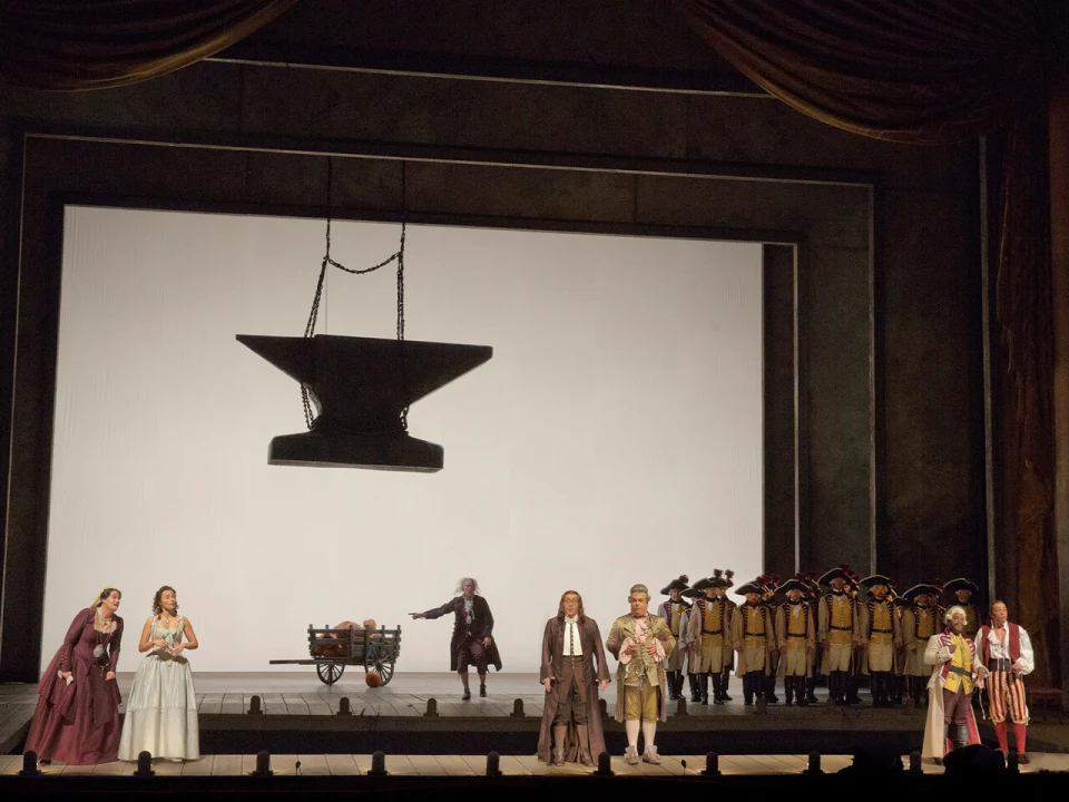 Stage production scene featuring performers in historical costumes, a large suspended anvil, and a group of soldiers in the background. A wagon is also seen onstage.