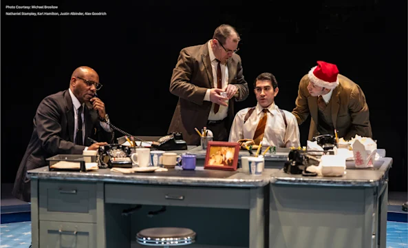 Four men in business attire are gathered around a cluttered office desk; one wears a Santa hat. Office supplies, phones, food containers, and a framed photo are on the desk.