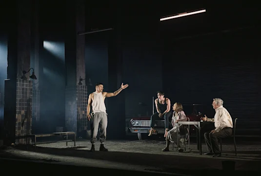 Four people perform on a dimly lit stage set to resemble a garage; one stands gesturing, one sits on a car, two sit at a table with drinks.