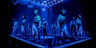 A group of shirtless male performers in jeans stand on a stage illuminated by blue lights in a nightclub or theater setting.