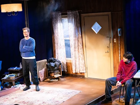 Two people stand and sit in a cluttered living room with blue walls and a beige door. One person stands with arms crossed, while the other sits on a chair near the door.