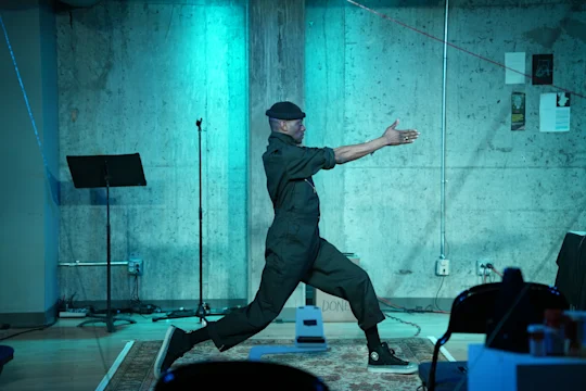 A person in dark clothing and a cap performs a dramatic pose with arms extended in a room with blue lighting and concrete walls.