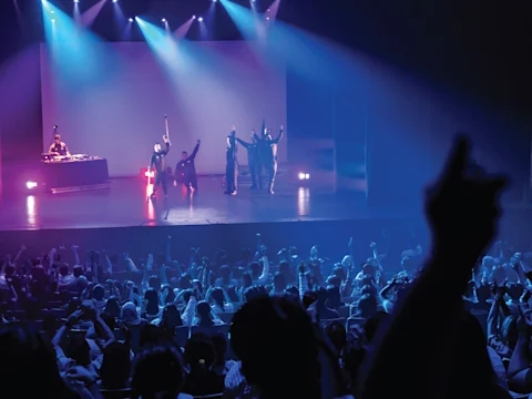 A crowd watches performers on stage with raised arms, illuminated by blue and purple lighting, while a DJ stands at a table to the left.