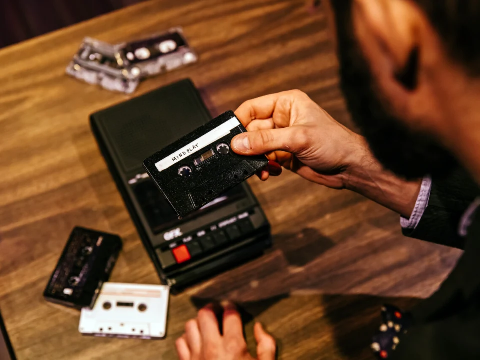 A person holding a cassette tape labeled "Mix Tape" next to a cassette player and other tapes on a wooden table.