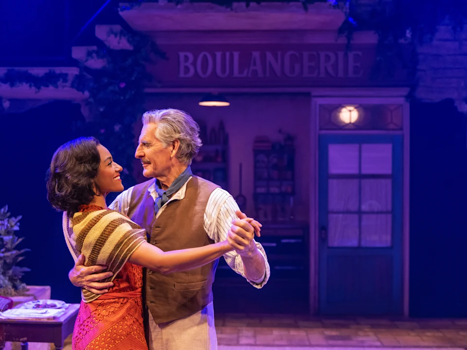 Production photo of The Baker's Wife in New York, showing a man and woman dance together in front of a storefront with a sign reading "Boulangerie" in a warmly lit stage setting.