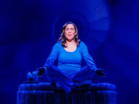 A woman in blue clothing sits cross-legged on a cushion with open arms, illuminated by blue stage lighting, against a dark backdrop.