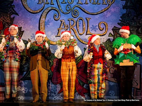 Five actors in colorful costumes and Santa hats stand onstage holding festive garlands and decorations in front of a backdrop reading "A Christmas Carol.