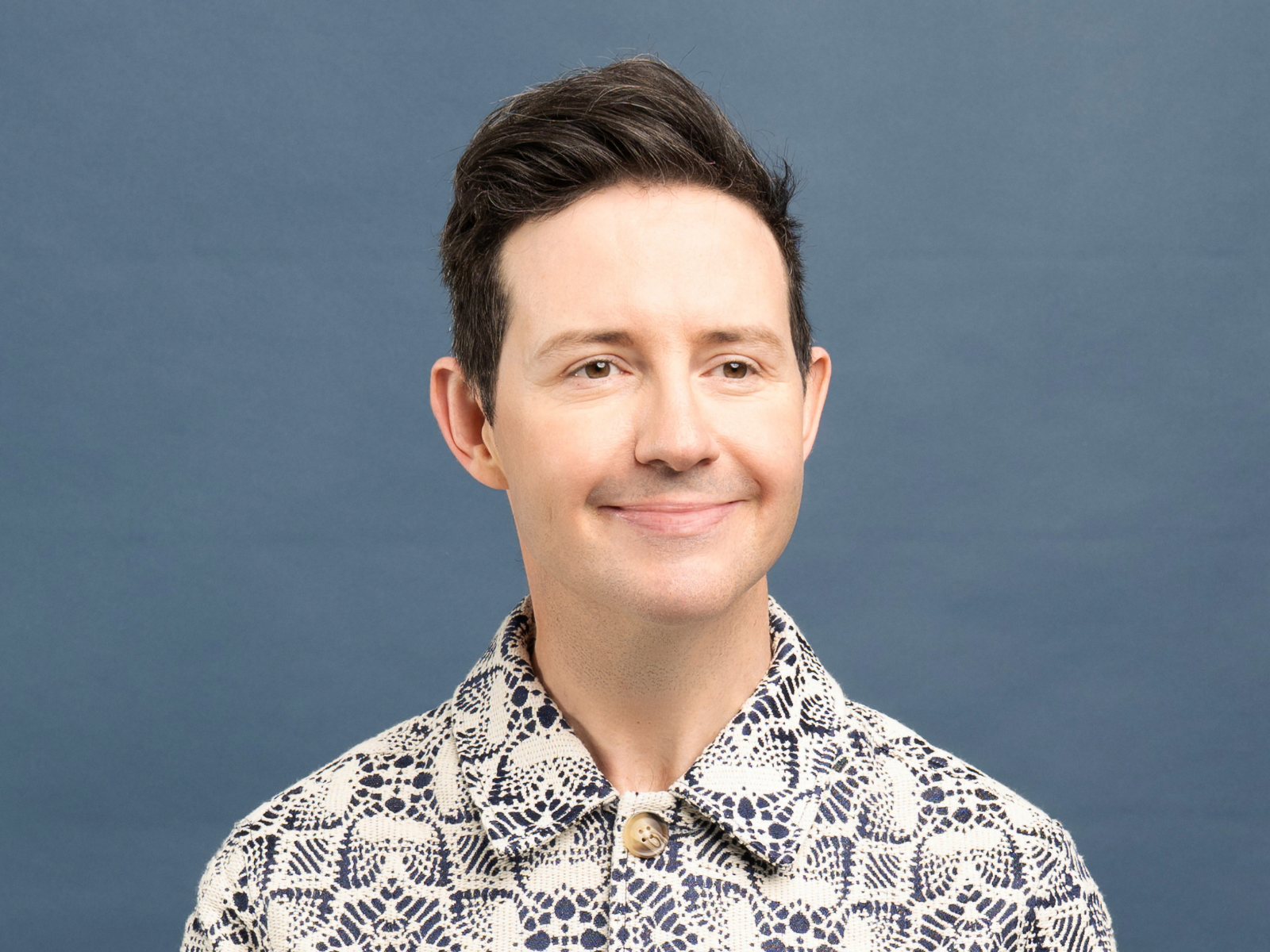 A person with short dark hair smiles slightly, wearing a patterned button-up shirt, in front of a plain blue background.