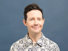 A person with short dark hair smiles slightly, wearing a patterned button-up shirt, in front of a plain blue background.