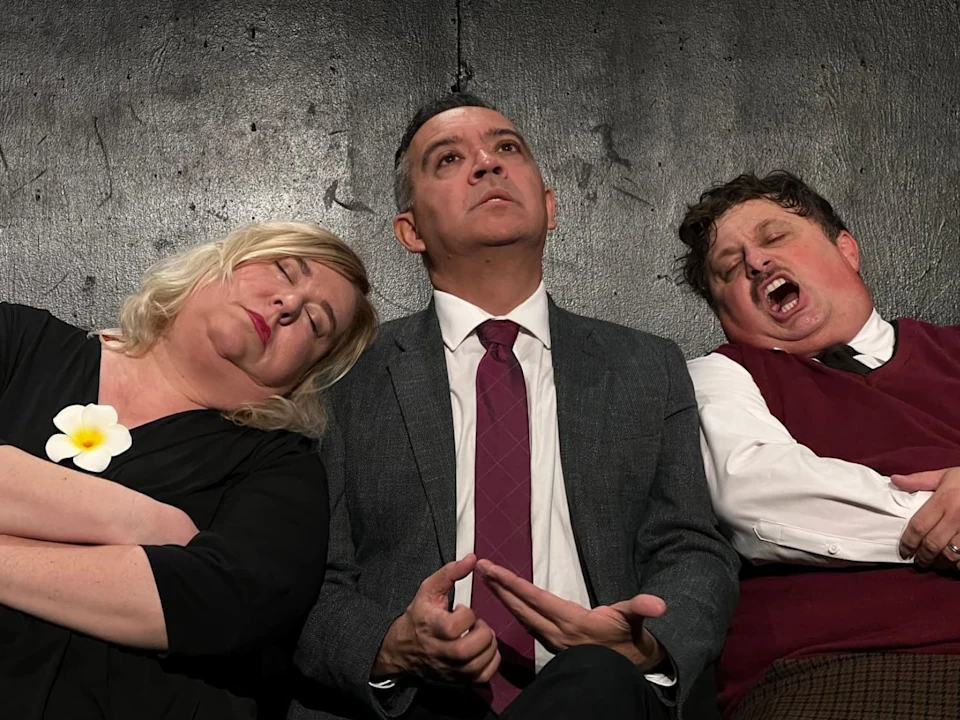 Three people sit against a dark wall; the two on either side appear to be dozing off while the person in the center looks upward with an exasperated expression.