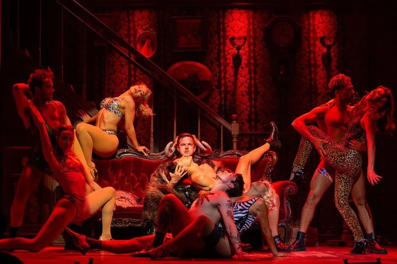 Performers in revealing, animal-themed costumes, pose dramatically on and around a vintage sofa, set against a richly decorated room with red lighting and ornate furnishings.