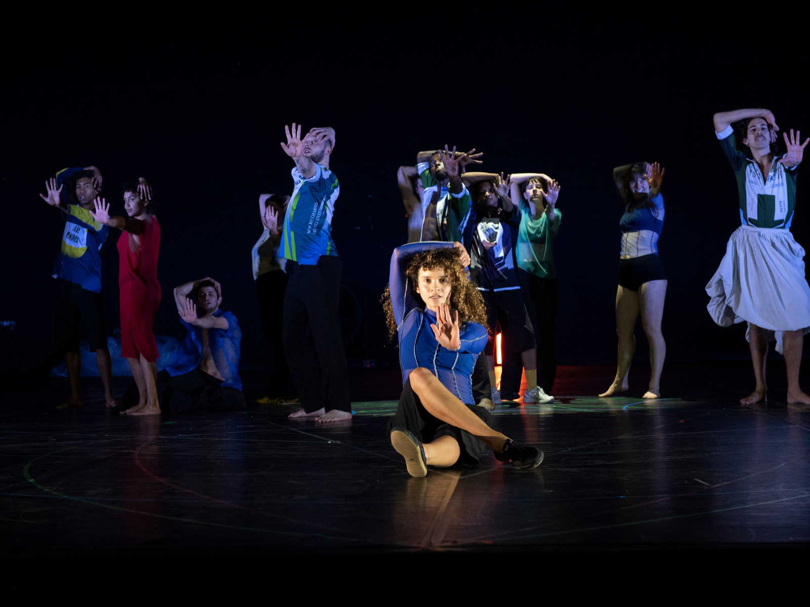 A group of dancers perform on stage, striking expressive poses with outstretched arms; one dancer sits in front with a raised hand.