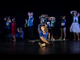 A group of dancers perform on stage, striking expressive poses with outstretched arms; one dancer sits in front with a raised hand.