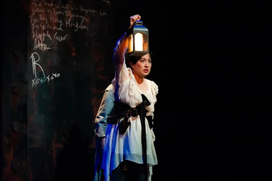 A woman in period costume holds a lit lantern aloft on a dark stage with chalk writing on a black wall behind her.