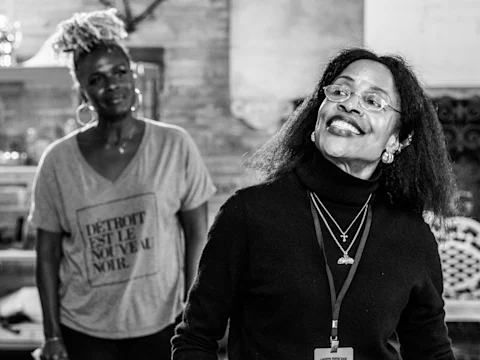 Two women indoors; one in the foreground smiling and wearing glasses and a lanyard, another in the background wearing a t-shirt. The scene is in black and white.