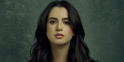 A woman with long, dark wavy hair and a neutral expression poses against a textured dark green background.