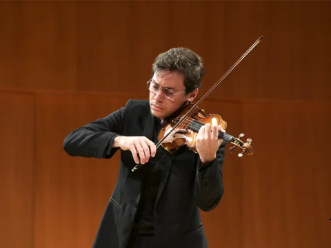 A person wearing glasses and a black suit plays a violin on a wooden stage in a focused manner.