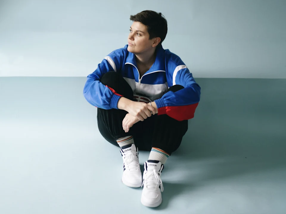 Person in a blue, white, and red jacket sits on the floor with knees up, looking to the side, wearing black pants and white sneakers against a light blue background.