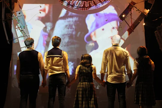 Five people in matching outfits stand holding hands, facing a large screen displaying a colorful, abstract projection in a dimly lit room.
