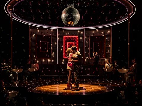 Two people embrace on a circular stage under a disco ball, surrounded by seated figures and illuminated by dramatic, colorful lighting.