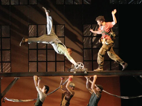 Performers balance on and support a metal beam during an acrobatic act on stage.