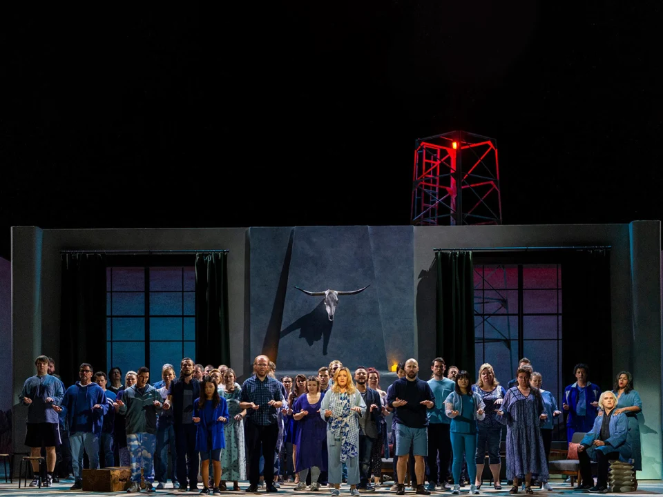 A group of performers in blue outfits stand on stage, with a bull head mounted on the wall behind them and a red-lit tower in the background.