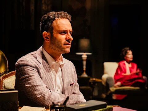A man in a light-colored suit sits at a desk looking serious, with a woman in red seated in the background. The setting appears to be a dimly lit room.