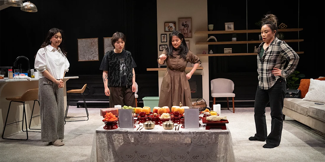 Four women stand around a table with food offerings in a modern living room, appearing focused and contemplative.
