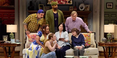 Seven people of various ages sit and stand around a living room couch, looking intently at a smartphone one woman is holding, with home decor visible in the background.