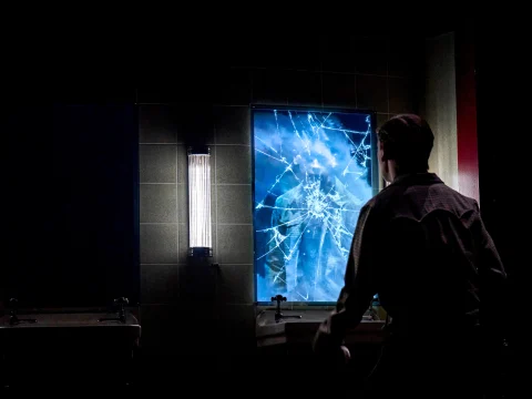 Louis McCartney as Henry Creel in Stranger Things: The First Shadow stands in front of a shattered, blue-lit mirror, their reflection fragmented by the cracks.