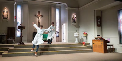 Two people in white robes walk down the steps of a church altar; one appears to be slipping or losing balance. Religious statues and a crucifix are visible in the background.