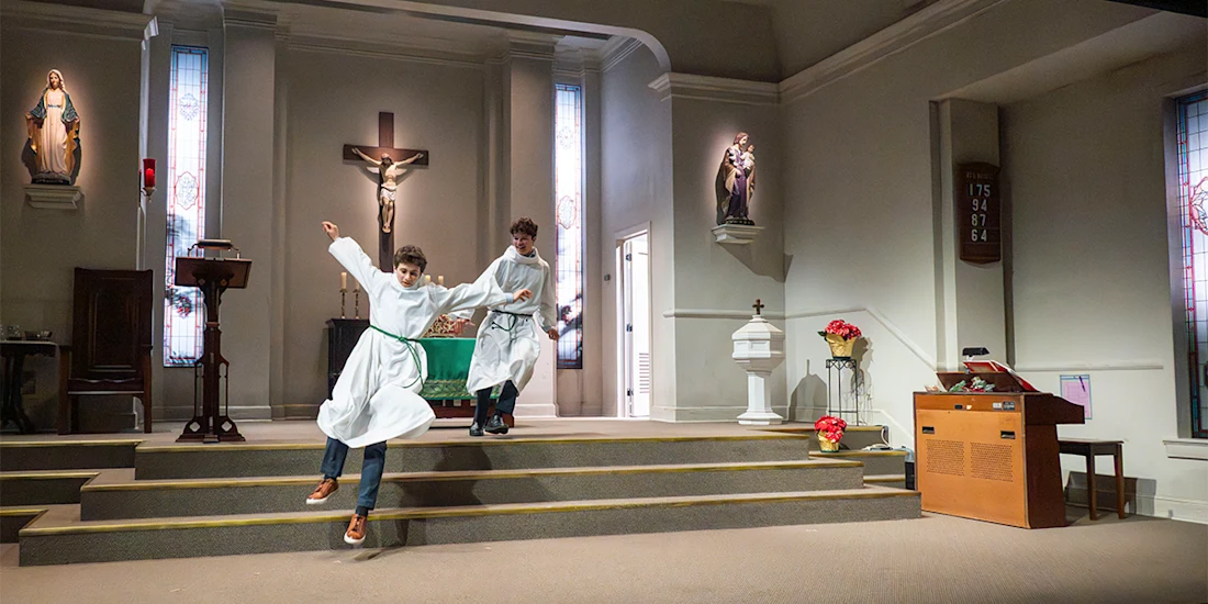 Two people in white robes walk down the steps of a church altar; one appears to be slipping or losing balance. Religious statues and a crucifix are visible in the background.