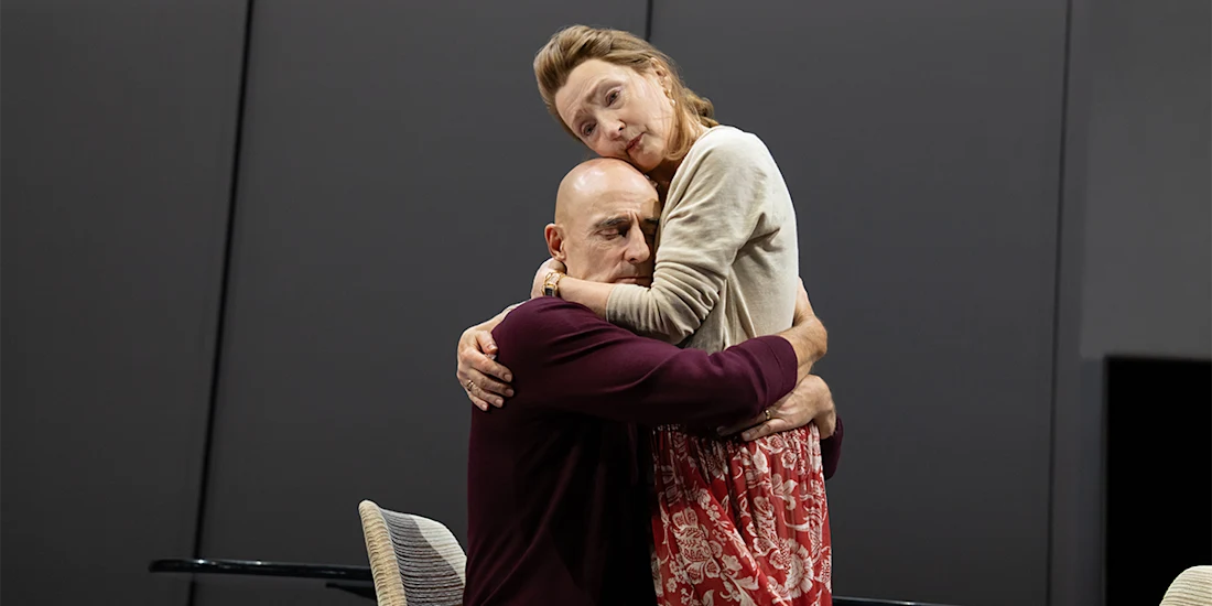 A bald man sitting embraces a standing woman in a red skirt and beige sweater against a plain, dark background.
