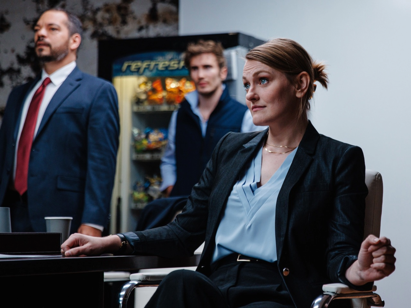 Three people in business attire are in an office; a woman sits at a table while two men stand behind her. A vending machine with snacks is visible in the background.