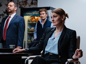 Three people in business attire are in an office; a woman sits at a table while two men stand behind her. A vending machine with snacks is visible in the background.
