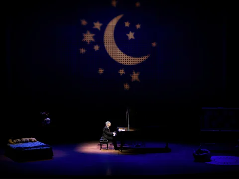 A person plays a grand piano on a dimly lit stage, with a bed and other furniture nearby and a crescent moon and stars projected on the backdrop.
