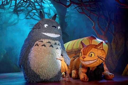 A person in a yellow dress stands beside large Totoro and Catbus costumes on a stage set with trees and blue lighting, evoking a scene from "My Neighbor Totoro.