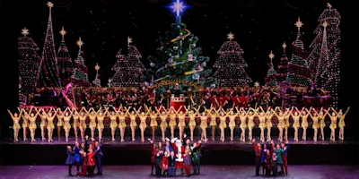 A large group of dancers in gold costumes perform in front of an orchestra and decorated Christmas tree, with festive holiday lights in the background.