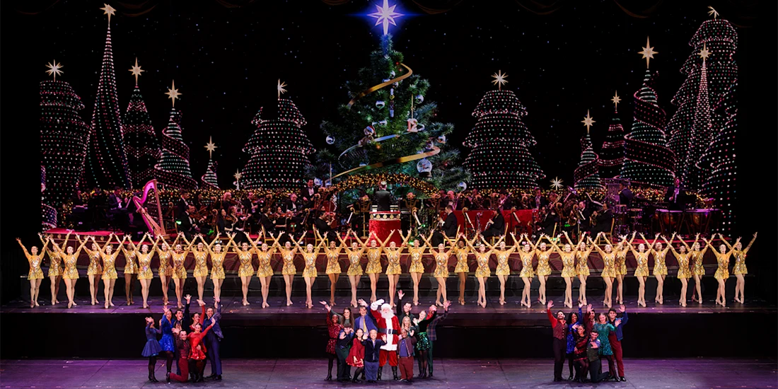 A large group of dancers in gold costumes perform in front of an orchestra and decorated Christmas tree, with festive holiday lights in the background.